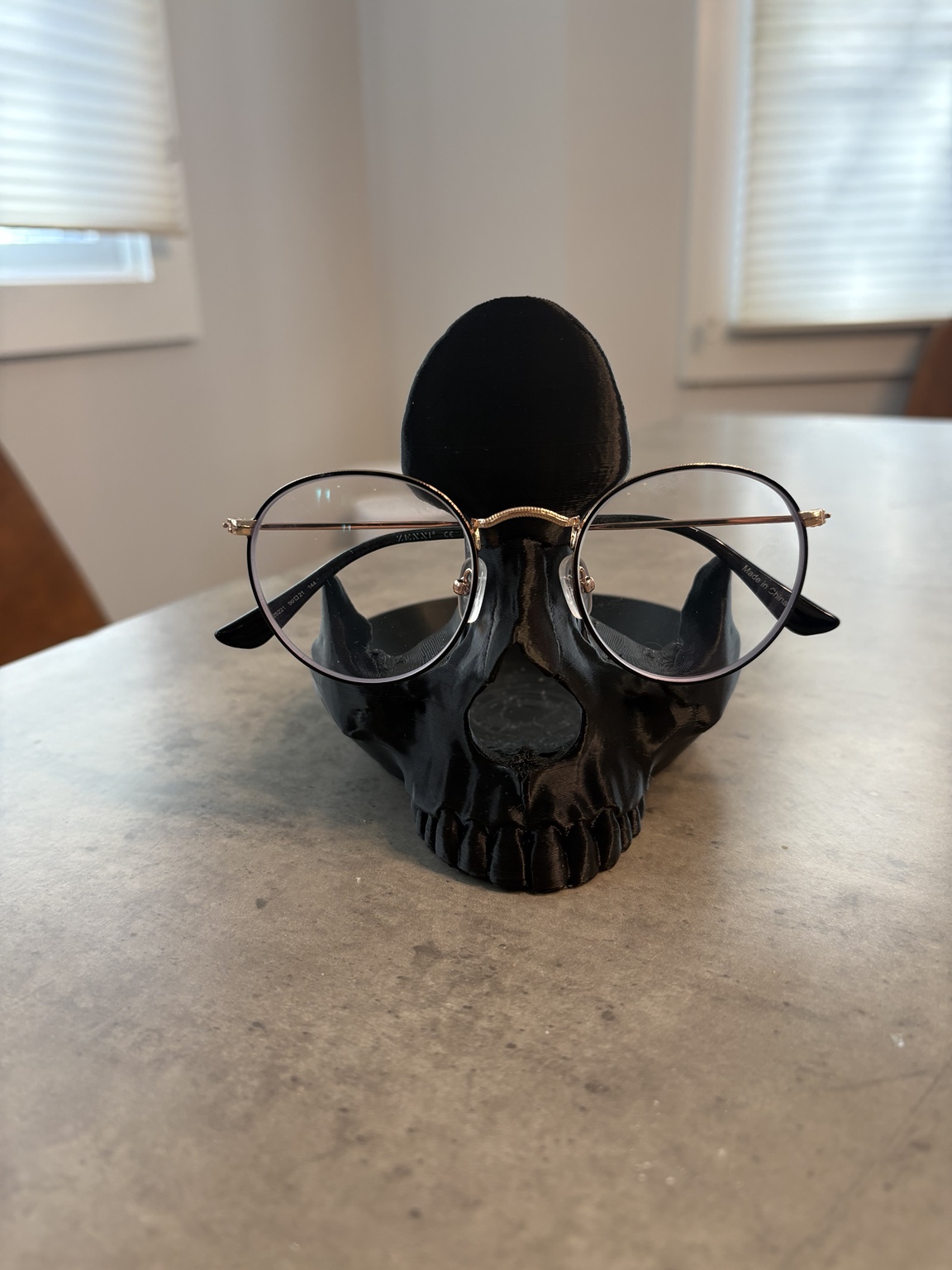 Black 3D-printed skull holding a pair of round eyeglasses on a table.