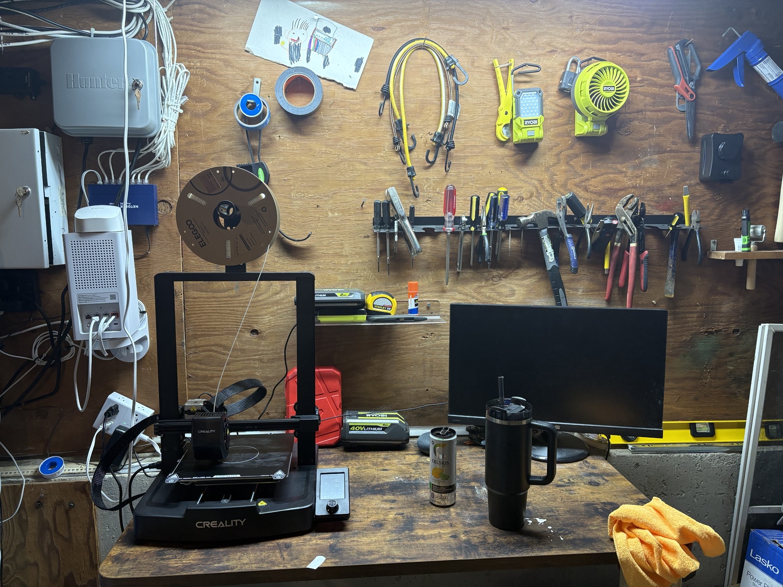 Creality Ender-3 printer on a workbench beneath hanging tools, beside a monitor and filament spool.